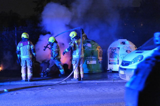 Feuerwehrleute in Helmen stehen auf einer Straße mit einem brennenden Auto, Rauchwolken, Bäumen und einem Zaun im Hintergrund.