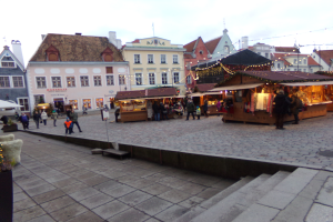 Ein geschäftiger Weihnachtsmarkt in Tallinn, Estland mit Menschen um dekorierte Stände, festliche Lichter, Gebäude im Hintergrund, bewölkter Himmel, Treppe nach oben und Topfpflanzen.