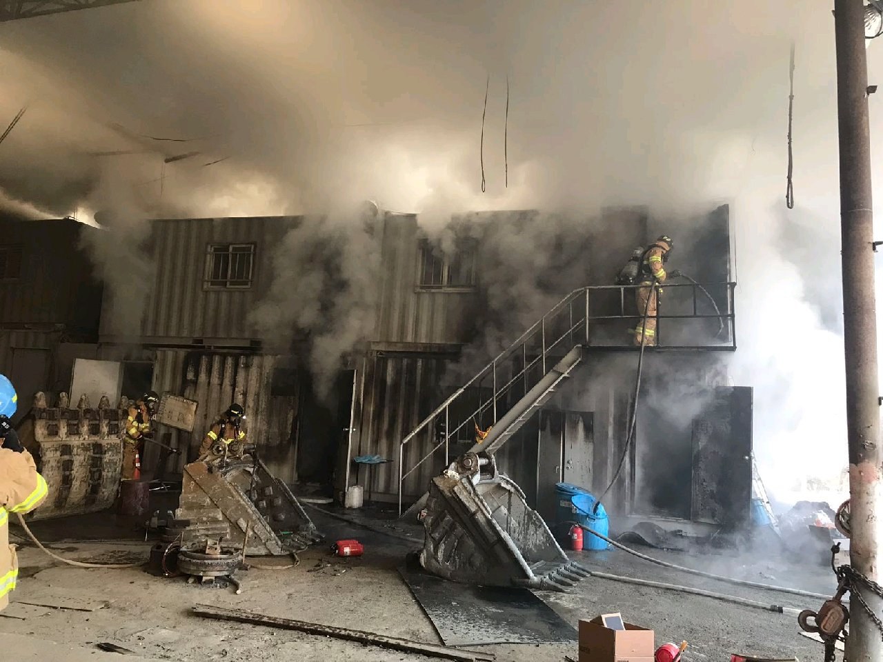 Feuerwehrleute in Helmen und Schutzausrüstung arbeiten daran, ein Lagerfeuer zu löschen, mit Rauch, der aus dem Gebäude aufsteigt, und Trümmern, die auf dem Boden verstreut sind.