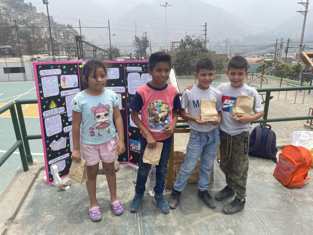 Gruppe von Kindern auf einem Basketballfeld mit Papiertüten in den Händen, mit Vorräten auf dem Boden, einer Tafel mit Text auf der linken Seite und einem Zaun, Pfosten und Gebäuden im Hintergrund.