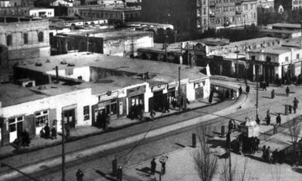 Schwarzes und weißes Foto einer Stadtstraße mit Fußgängern, Gebäuden, Bäumen, Strommasten und Drähten, das die historische Architektur und Stadtplanung der Stadt widerspiegelt.