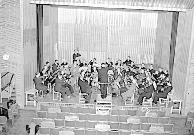 Schwarzes und weißes Foto eines großen Orchesters, das auf einer Bühne in einem großen Auditorium spielt, mit einer Uhr an der linken Wand und Reihen leerer Stühle unten.