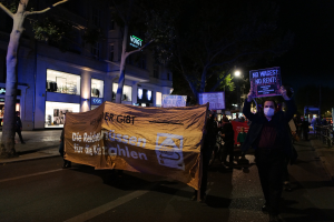 Gruppe von Menschen marschiert nachts in Berlin, hält Protestschilder, mit Bäumen, Gebäuden, Fahrrädern und Laternen im Hintergrund.