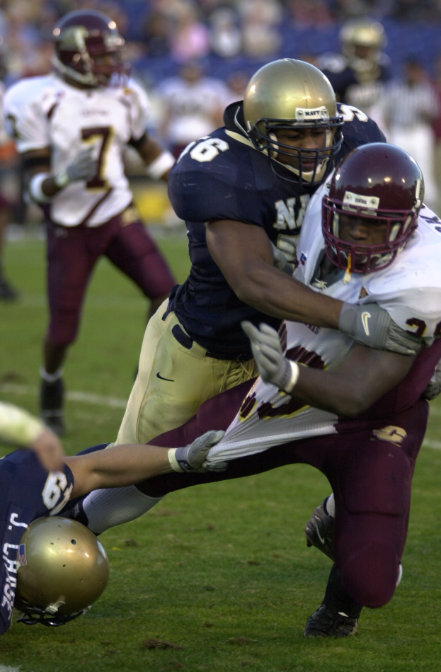 Zwei Fussballspieler in Helmen und Uniformen räumen sich auf dem Feld, während andere im Hintergrund zuschauen.