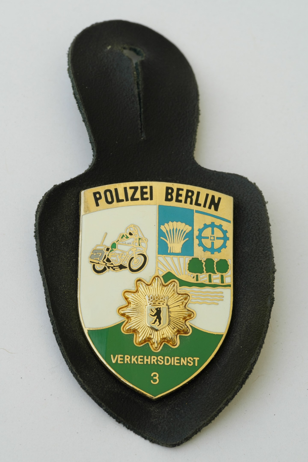 Ein schildförmiges Polizeibadge mit 'Polizei Berlin' in schwarzer Schrift, das einen blauen Hintergrund, einen weißen Stern und einen weißen Rand hat und auf einer Oberfläche liegt.