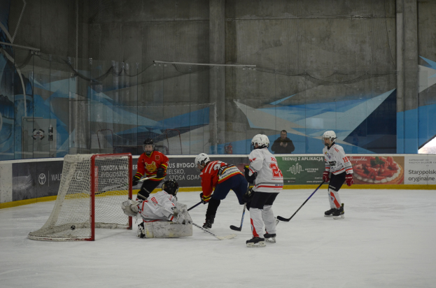 Gruppe von Menschen in Helmen und Eishockeyausrüstung beim Eishockeyspielen auf einer Indoor-Eisbahn mit einem Tor auf der linken Seite und Bannern im Hintergrund.