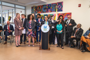 Gruppe von Menschen vor einem Podium mit Mikrofon und einer Tafel mit der Aufschrift "Sancho Stadtrat tritt dem Stadtrat bei", mit sitzenden und stehenden Teilnehmern, Flaggen, Feuerlöscher und Deckenleuchten im Hintergrund.