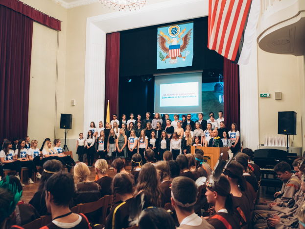 Eine Gruppe von Menschen steht an einem Podium mit einem Mikrofon, flankiert von Fahnen und Lautsprechern, und spricht eine sitzende Menge bei einem kulturellen Ereignis an.