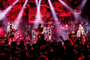 Ein Live-Auftritt einer japanischen Rockband mit Musikern, die Gitarren spielen und singen, auf der Bühne, begleitet von Mikrofonen und Ständern, während das Publikum unten zuschaut.