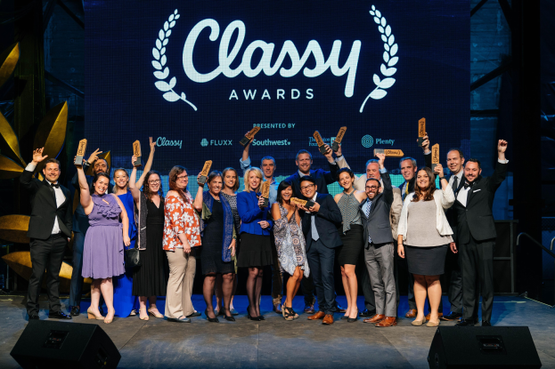 Gruppe von Menschen auf einer Bühne mit Auszeichnungen, darunter Lautsprecher und ein Bildschirm mit der Aufschrift "Classy Awards 2019" im Hintergrund.