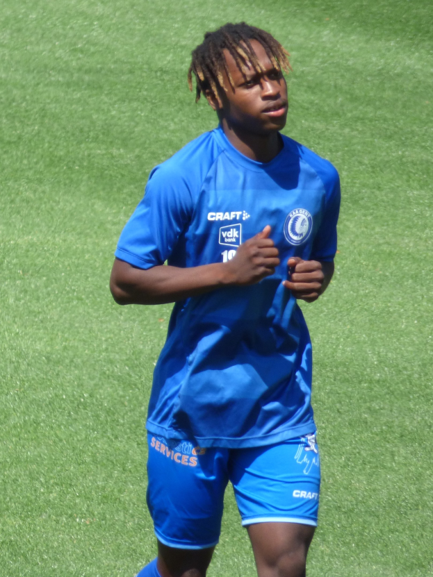 Ein junger Mann in einem blauen T-Shirt und Shorts rennt auf einem Fußballfeld mit ausgestreckten Armen während eines Spiels.