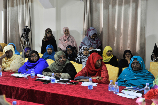 Eine Gruppe von Frauen, die an einem Tisch sitzen und verschiedene Gegenstände haben, diskutieren mit einer Vase auf der rechten Seite und Vorhängen im Hintergrund.