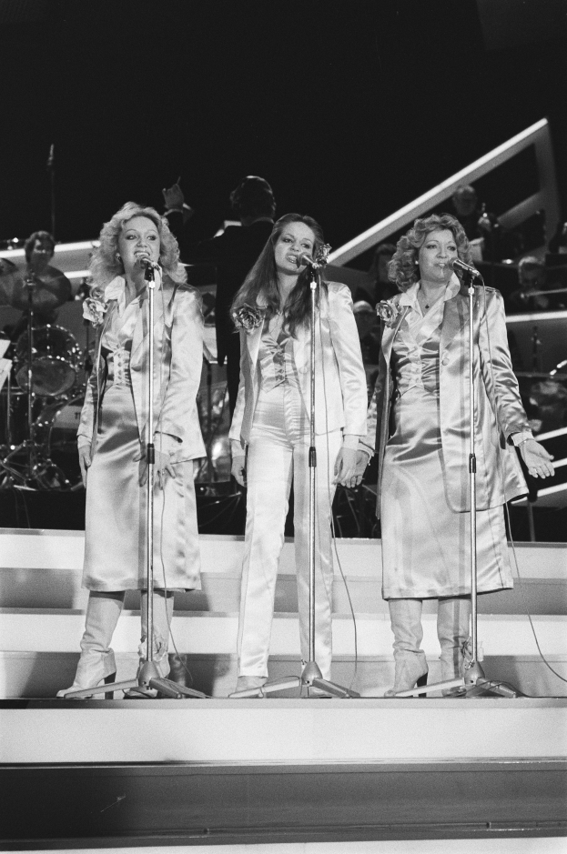 Drei Frauen singen in ein Mikrofon auf der Bühne mit Musikern, die hinter ihnen spielen.