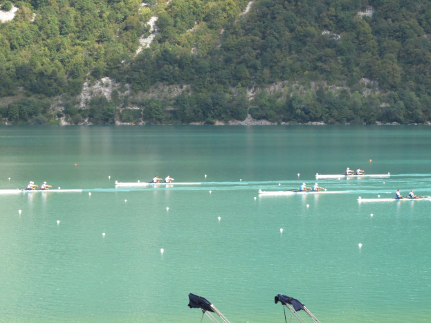 Gruppe von Menschen beim Ruderwettbewerb auf einem Gewässer mit Bäumen und einem Hügel im Hintergrund, Kleidung über Stühle am Wasser gelegt.