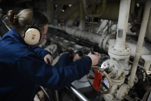 Zwei Frauen in blauen Uniformen arbeiten an einer Maschine in einer Öl- und Gasindustrieumgebung, wobei eine ein Objekt hält und Kopfhörer trägt, und im Hintergrund Rohre sichtbar sind.