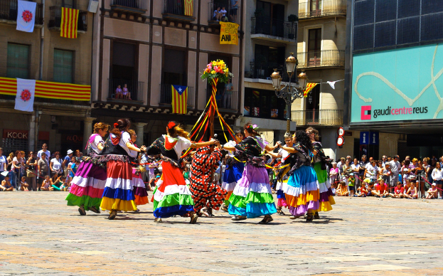 Eine Gruppe von Frauen in bunten Kleidern tanzt auf der Straße, umgeben von einer Menge Menschen, mit Gebäuden, Geländern, Balkonen, Fahnen, einem Laternenmast und einer Tafel mit Schrift im Hintergrund.