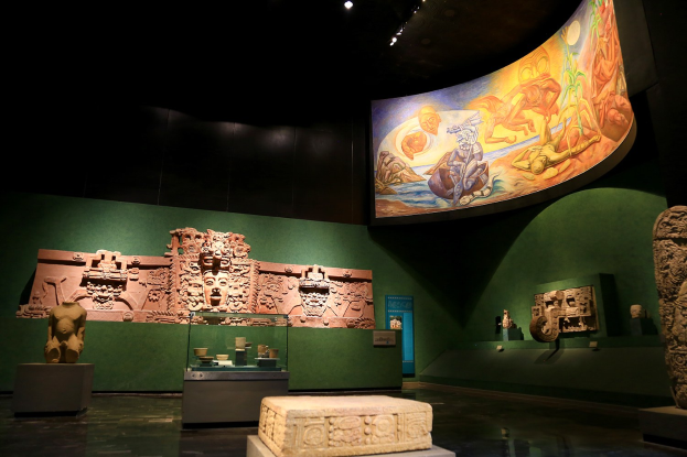 Ausstellungsraum im Anthropologischen Museum von Mexiko-Stadt mit verschiedenen Kunstwerken, darunter Skulpturen, Tischgegenstände, eine Texttafel, eine Wandmalerei und Deckenbeleuchtung.