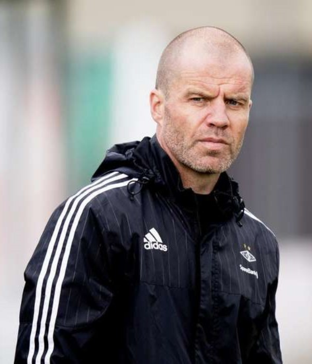 Bald kahlköpfiger Trainer in marineblauer Jacke mit weißen Streifen und weißem Kragen auf einem Fußballfeld stehend