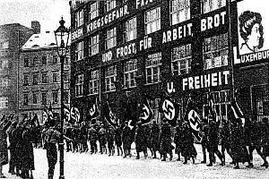 Schwarzes und weißes Bild einer Gruppe, die mit Fahnen und Transparenten die Straße entlangmarschiert, mit einem Lichtmast links und einem Gebäude mit einem Nazi-Parteischild im Hintergrund.