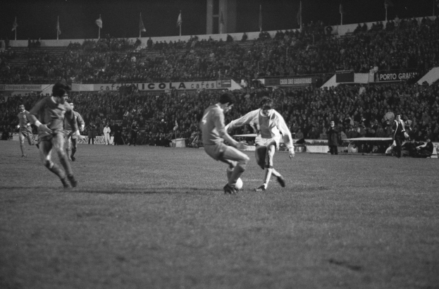 Schwarzes und weißes Bild von Männern, die Fußball auf einem Feld spielen, mit Zuschauern auf den Tribünen und Fahnen im Hintergrund.