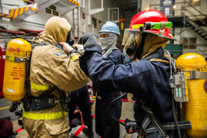 Eine Gruppe von Feuerwehrleuten in Schutzausrüstung, einschließlich Helmen, Handschuhen und Sauerstoffflaschen, steht zusammen mit Taschen auf dem Boden, mit Treppen, Geländern, Lampen und Brettern im Hintergrund.