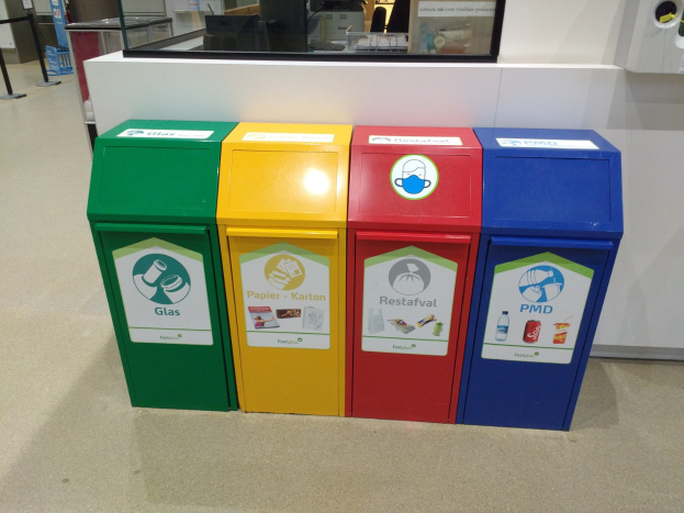 Vier farbenfrohe Mülltonnen in einer Reihe auf dem Boden, mit einer Arbeitsplatte, einem Monitor und anderen Gegenständen im Hintergrund und einer Person links daneben.