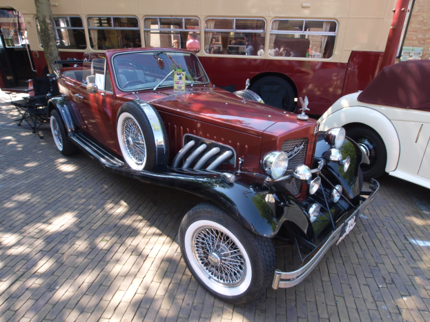 Vintage-Auto neben einem Doppeldeckerbus auf einer Kopfsteinpflasterstraße geparkt, mit Stühlen und einem Baum im Vordergrund, einem Gebäude mit Fenstern im Hintergrund und Menschen im Bus sichtbar.