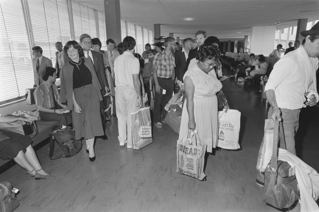 Ein Schwarz-Weiß-Bild von Reisenden in einem Flughafen, einige sitzen auf Bänken und andere stehen mit Gepäck, mit einem Mann und einer Frau, die Taschen halten, in der Mitte und Fensterläden links.