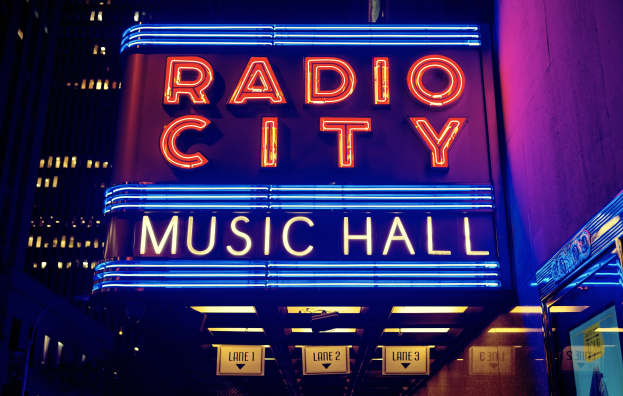 Radio City Music Hall in New York City, hell erleuchtet in der Nacht, umgeben von hohen Gebäuden, mit einem "Radio City"-Schild und zusätzlichen Schildern auf der rechten Seite.