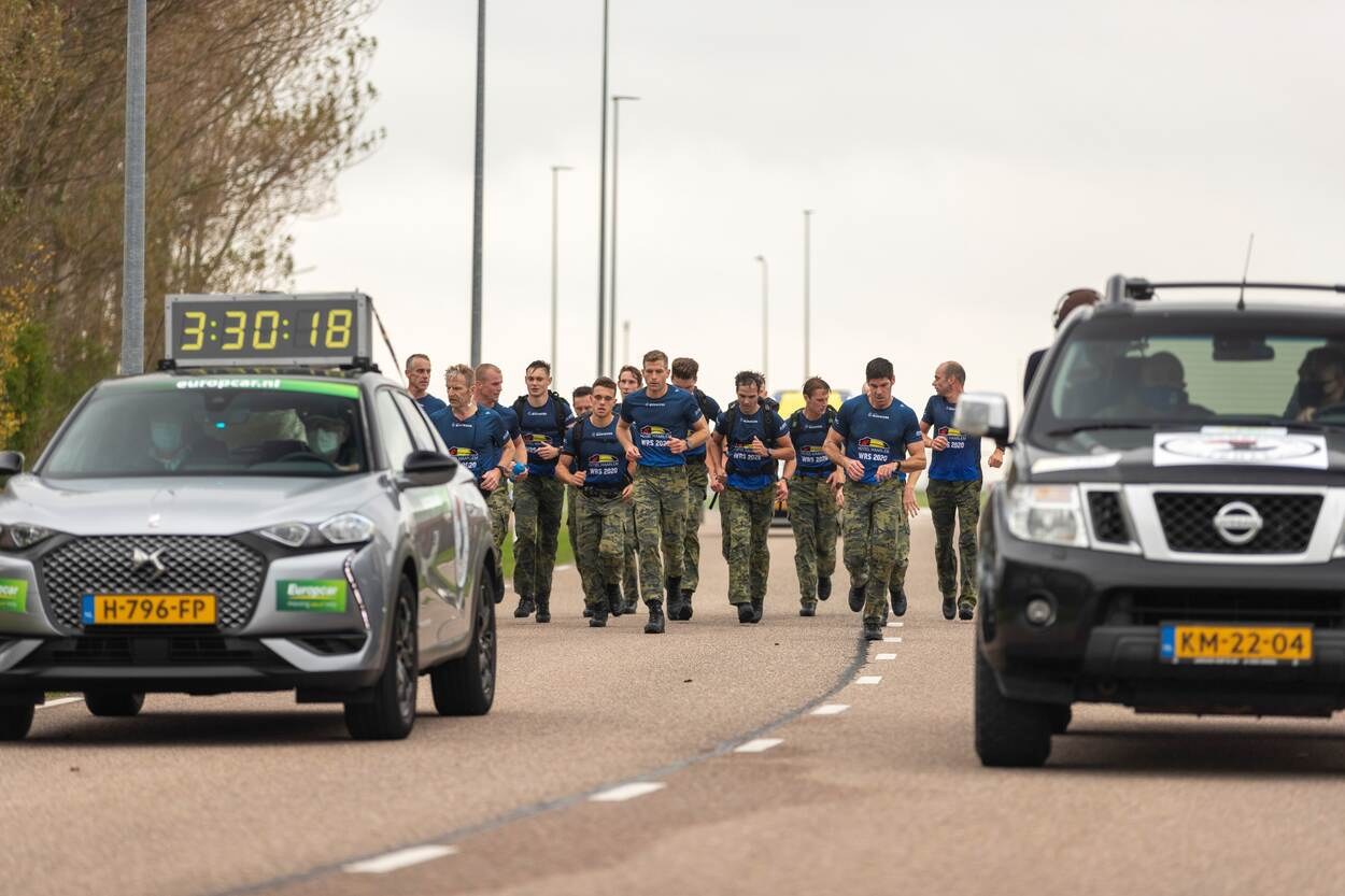 Eine Gruppe von Menschen, die auf einer Straße neben geparkten Autos laufen, mit Bäumen auf der linken Seite und Masten im Hintergrund, unter einem sichtbaren Himmel, bei einem Europäischen Cross-Country-Challenge.