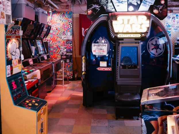 Ein hell erleuchteter Arcade-Raum mit mehreren Maschinen, darunter ein Star Wars Battle Pod, bunten Postern an den Wänden und verstreuten Gegenständen, die eine lebendige Atmosphäre schaffen.
