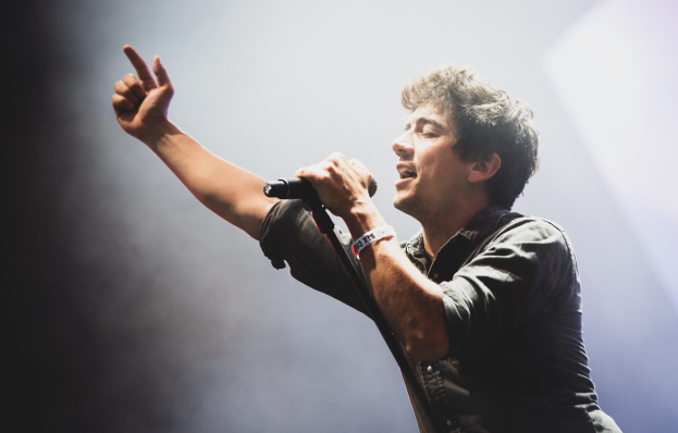 Ein Mann in schwarzem Hemd und Jeans singt leidenschaftlich in ein Mikrofon auf einer hell erleuchteten Bühne, die Augen geschlossen und das Haar stylisch zerzaust, während das Publikum im Hintergrund applaudiert.