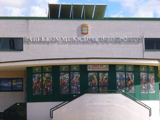 Ein Gebäude mit einer Tafel, die "Pabellón Municipal de Deportes" lautet, mit Fenstern, einer Treppe, einem Metallzaun, Pflanzen und Bäumen unter einem bewölkten Himmel.