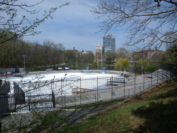 Eisstadion umgeben von einem Metallzaun, Gras, Bäumen, Fahrzeugen, Straßenlaternen, einem Gewässer, Gebäuden und einer bewölkten Himmel.