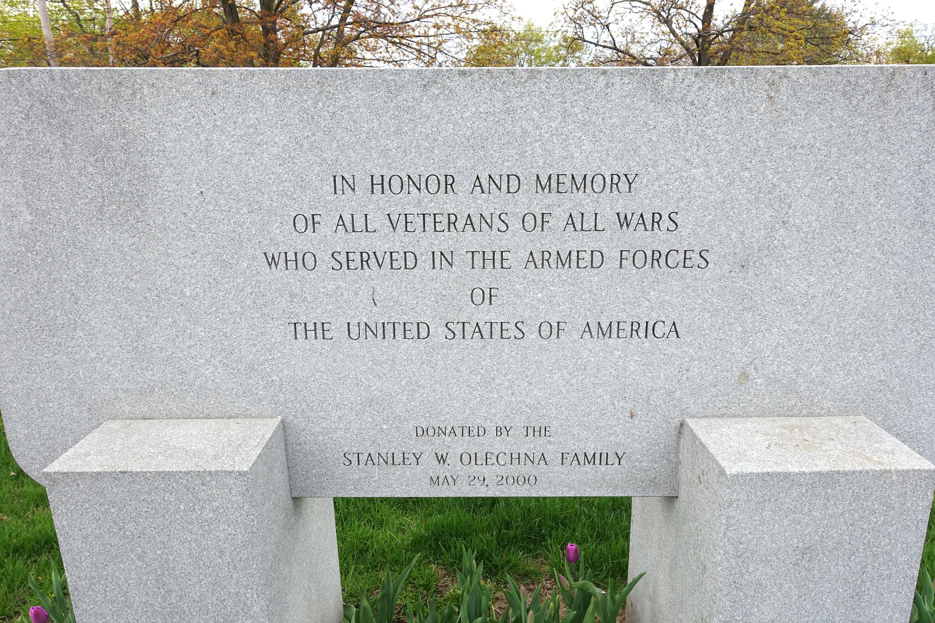 Gedenkstein mit Inschrift für alle US-Veteranen, umgeben von Pflanzen, Blumen, Gras, Bäumen und einer bewölkten Himmel