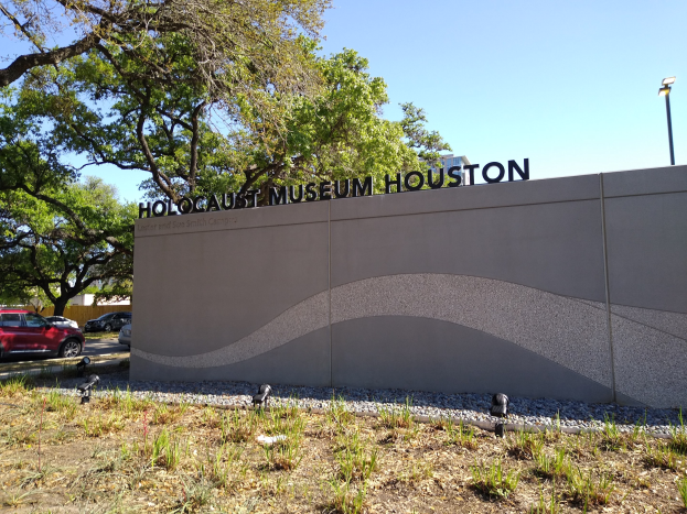 Außenansicht des Holocaust-Museums Houston mit einer Textwand, Gedenksteinen, Grünfläche, Bäumen, vorbeifahrenden Fahrzeugen, einer Straßenlaterne und einem bewölkten Himmel.