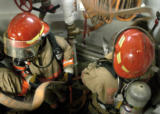 Feuerwehrleute in Helmen und Schutzausrüstung arbeiten an einem Feuerhydranten mit Rohren, Ventilen und einer Wand im Hintergrund, mit Text unten.