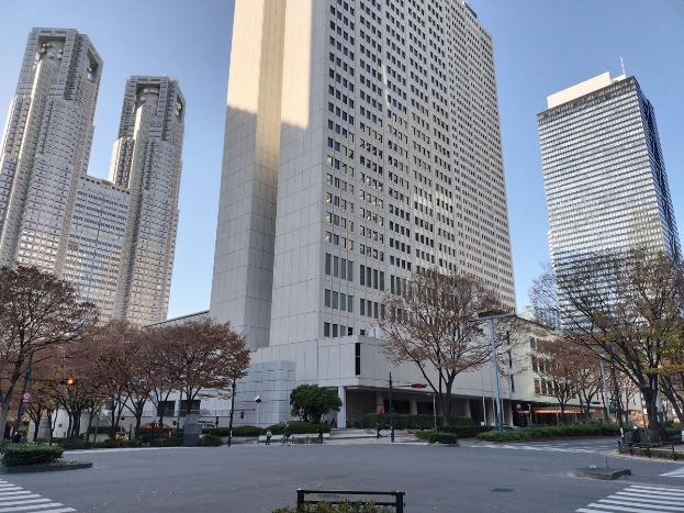 Eine Stadtstraße in Tokio, Japan, mit hohen Gebäuden, Bäumen, Laternen, Verkehrszeichen und einigen Menschen, wobei das Tokyo Metropolitan Government Building im Hintergrund zu sehen ist.