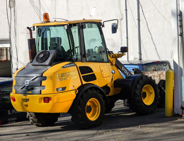 Gelber Radlader auf einer Straße vor einem Gebäude mit einer Wand im Hintergrund geparkt, mit anderen Fahrzeugen in der Nähe.