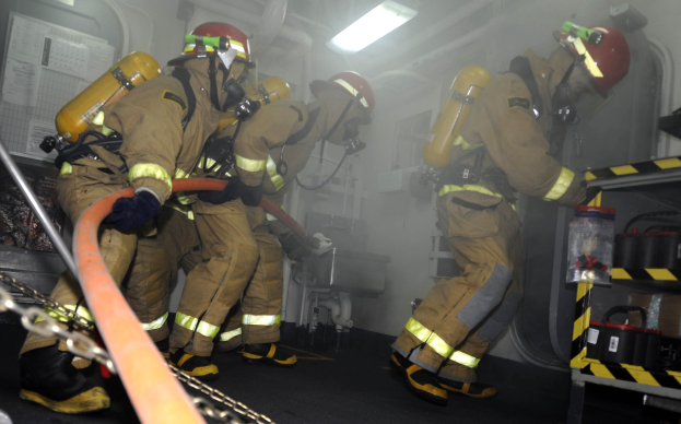 Feuerwehrleute in Schutzausrüstung arbeiten an einem Löschfahrzeug, mit Equipment-Racks und Hintergrund-Infrastruktur im Hintergrund.