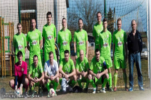 Eine Gruppe von Männern in grünen T-Shirts steht auf einem Fußballfeld, einige hocken, mit einem Ball vor ihnen in der Nähe eines Torpfostens, Bäume, Gebäude und Fahrzeuge im Hintergrund.