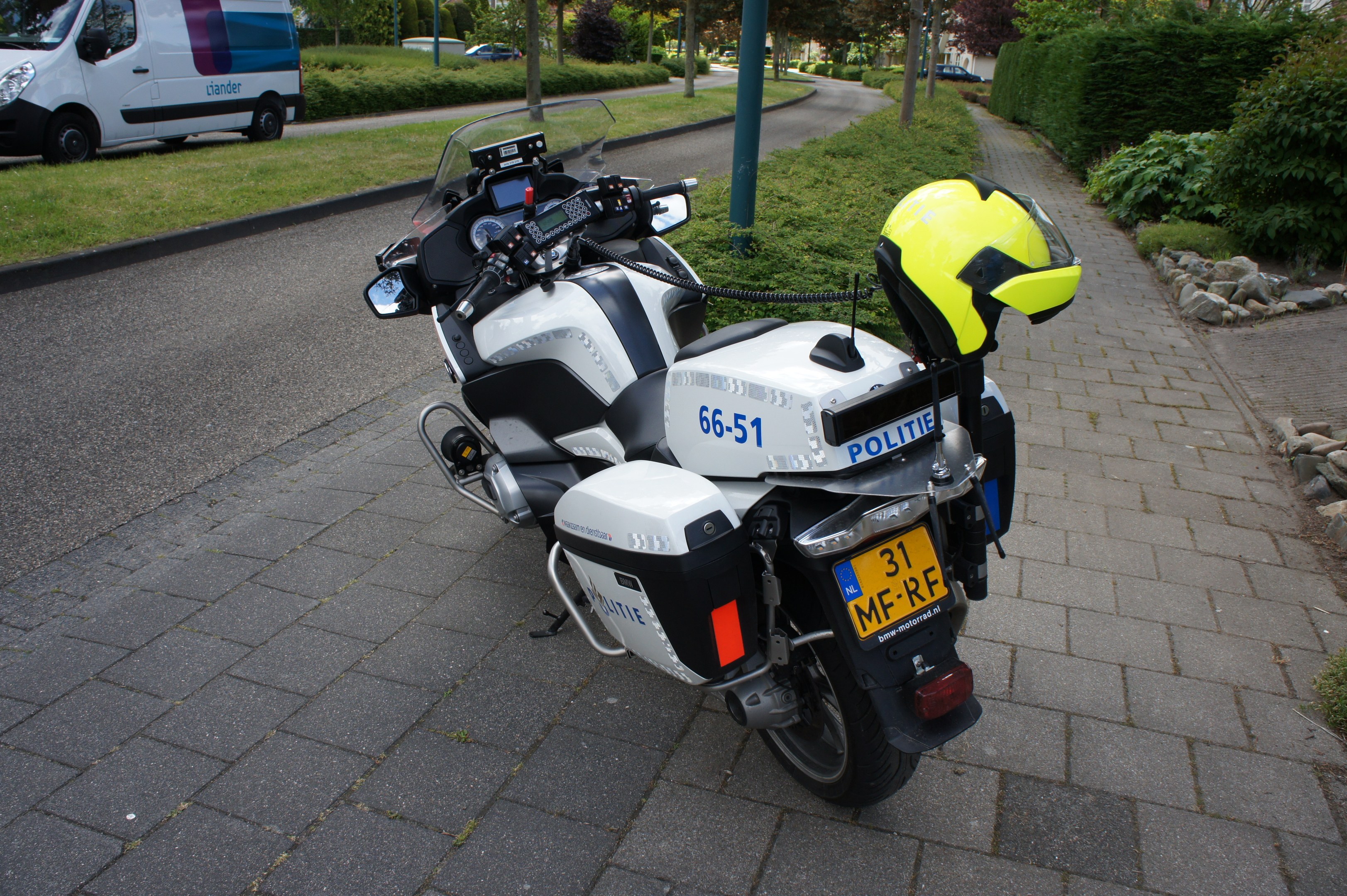 Ein Polizeimotorrad mit einem Helm darauf am Straßenrand geparkt, mit Bäumen, Pfählen, Gebäuden, Pflanzen, Gras, Steinen und anderen Fahrzeugen im Hintergrund.