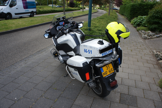 Ein Polizeimotorrad mit einem Helm darauf am Straßenrand geparkt, mit Bäumen, Pfählen, Gebäuden, Pflanzen, Gras, Steinen und anderen Fahrzeugen im Hintergrund.