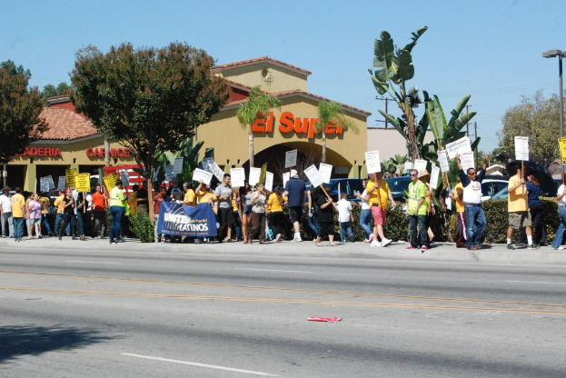 Menschen mit Schildern in einer Gruppe, mit kleinen Büschen, einer Fahne, einem Gebäude mit Namensschildern und Autos im Hintergrund.