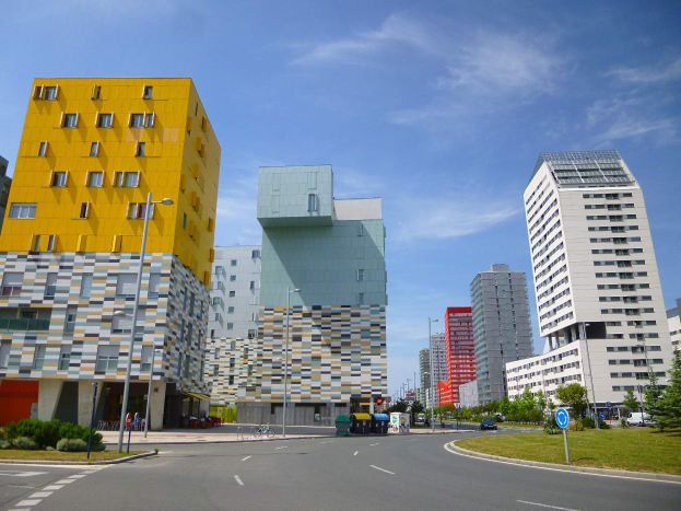Eine Stadtstraße mit hohen Gebäuden im Hintergrund, Straßenlaternen, Verkehrsschildern, Autos, Büschen, Bäumen und einer bewölkten Himmel, mit den einzigartig gestalteten Gebäuden der Universität Rotterdam.