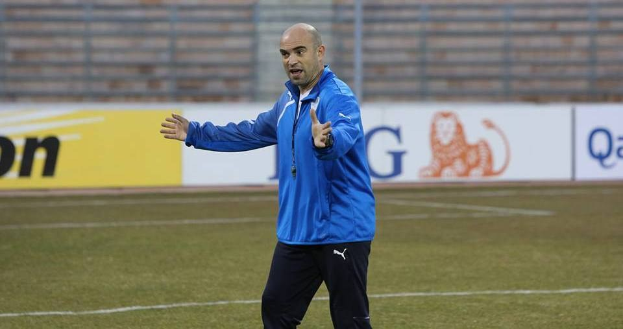 Ein Fußballtrainer in einer blauen Jacke steht entschlossen auf einem Feld, umgeben von Werbetafeln während eines Trainings.