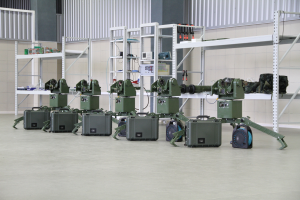 Eine Gruppe von Militärrobotern ist auf einem Boden umgeben von Koffern und anderen Gegenständen auf Gestellen positioniert, mit einer Wand und einem Geländer im Hintergrund.