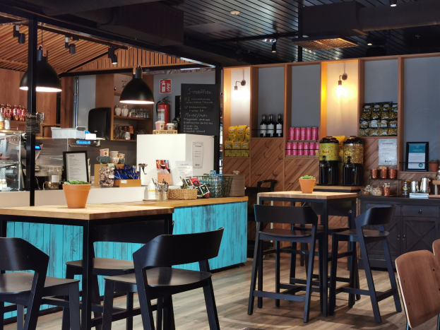 Innenraum eines Restaurants mit Holzmöbeln, einer Topfpflanze, einer Schiefertafel, einem Kühlschrank, einem Feuerlöscher, Regalen mit Flaschen, Schränken mit Gegenständen, einem Rahmenfoto an der Wand und Deckenlampen.