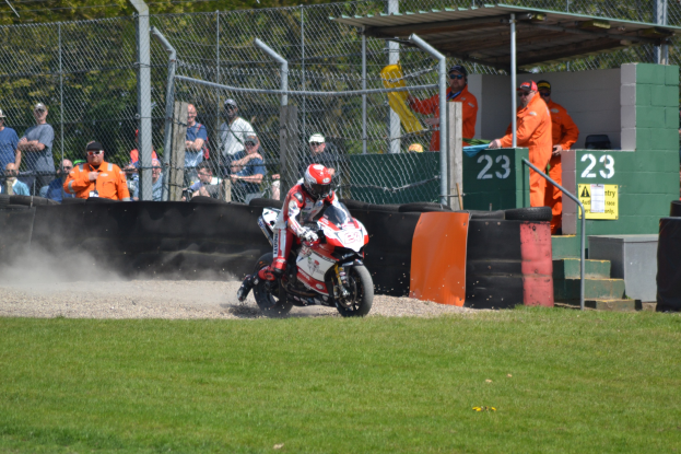 Ein Motorradfahrer in Helm und Handschuhen fährt über ein grasbewachsenes Feld, mit Zuschauern in Mützen und Schutzbrillen, einem Zaun, Treppen mit Geländern und einem nummerierten Schuppen im Hintergrund.