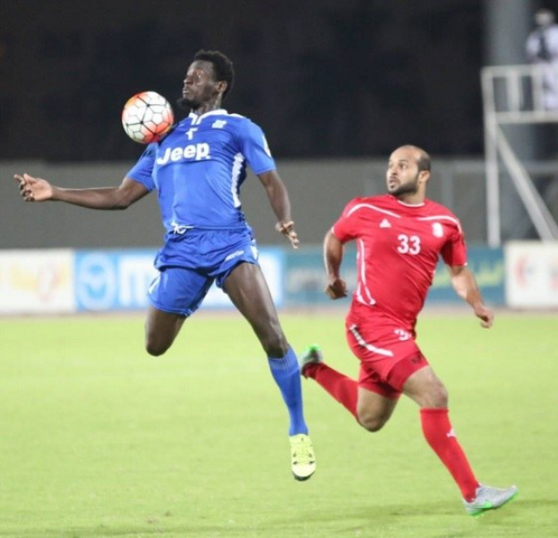 Zwei Männer beim Fußballspielen auf einem Feld, einer trägt ein blaues T-Shirt und Shorts, der andere ein rotes Kleid, im Hintergrund sind Bretter mit Text, ein Geländer, ein Pfahl und stehende Menschen zu sehen.